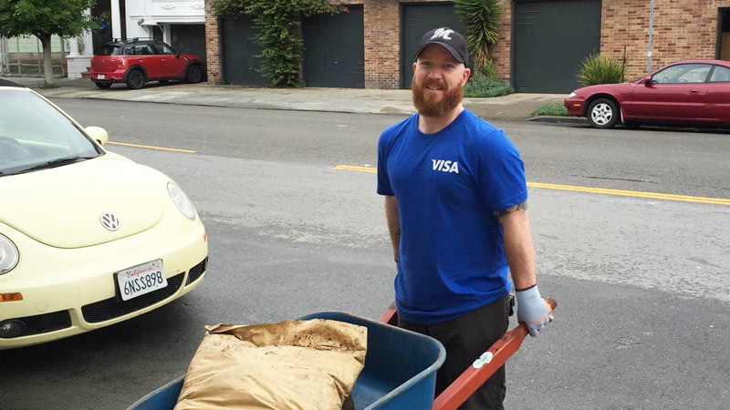 Visa volunteer using wheelbarrow full of landscaping supplies in urban environment.