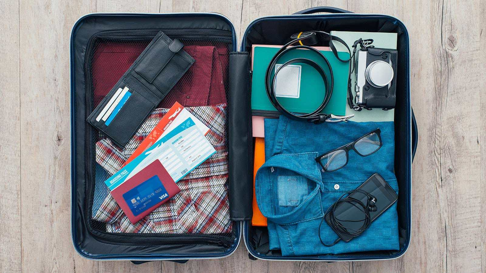 Open suitcase packed for travel with clothing, personal electronics, camera, wallet, airline tickets, passport and Visa card.