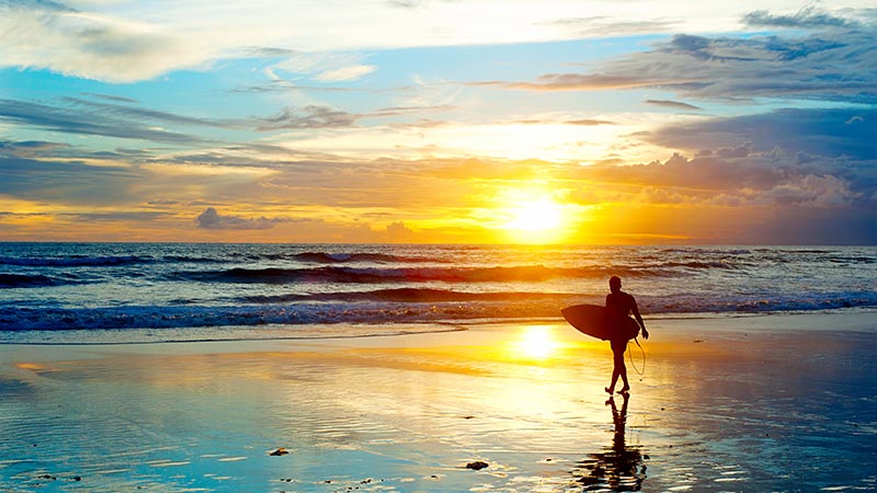 Gambar seorang pria bertualang di pantai dengan papan selancarnya menggunakan fasilitas biaya liburan ke Bali dari Visa